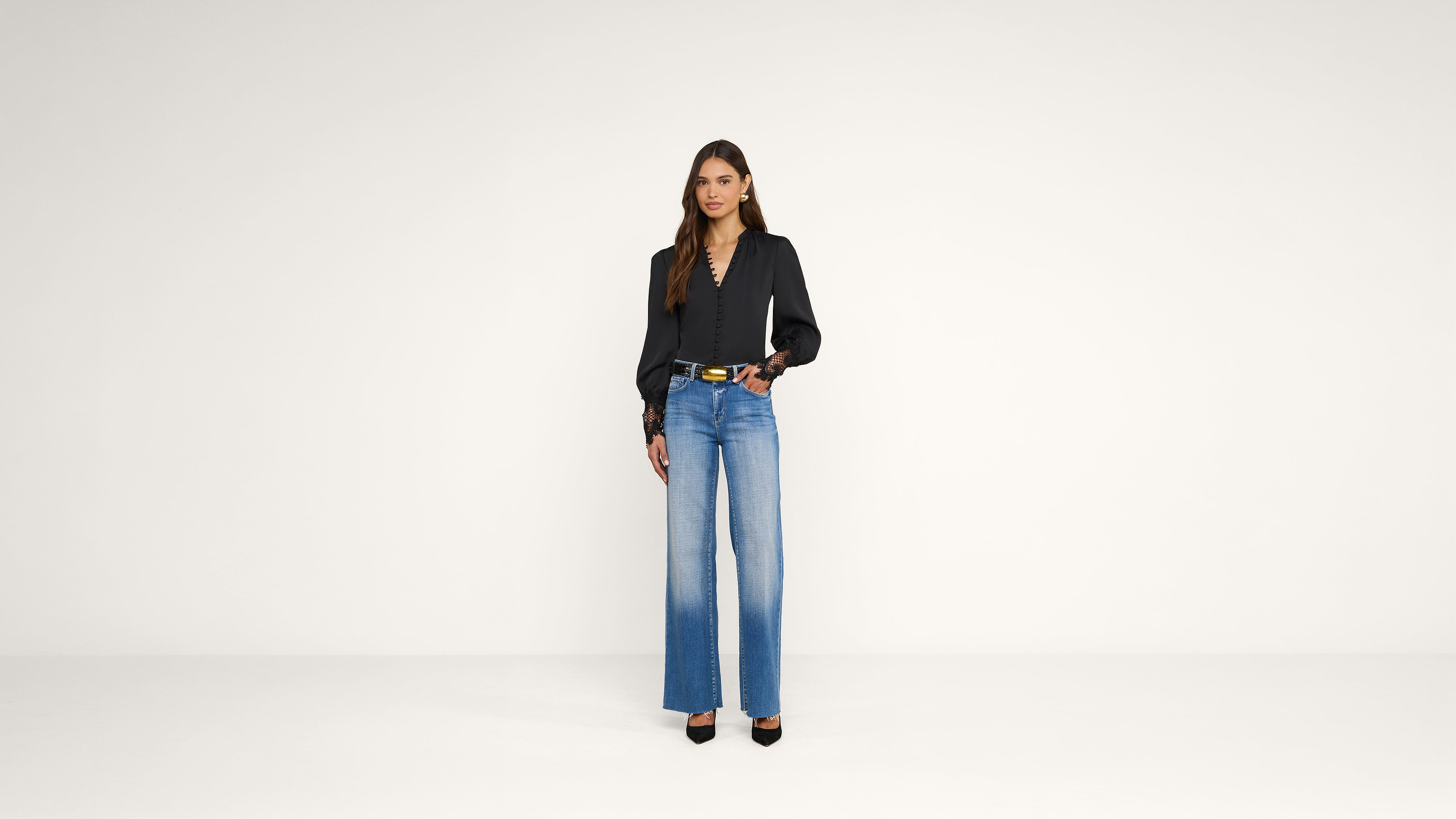 A woman stands against a plain white background, wearing a black long-sleeve top, blue wide-leg jeans, black heels, and a belt. She has long hair and is holding a phone in one hand.