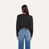Una donna è di spalle e indossa la camicetta Ava — un top nero di ispirazione vintage con polsini in pizzo guipure smerlato — abbinata a jeans blu a vita alta e una cintura nera. I suoi capelli castani lisci ricadono sciolti su uno sfondo beige a tinta unita.