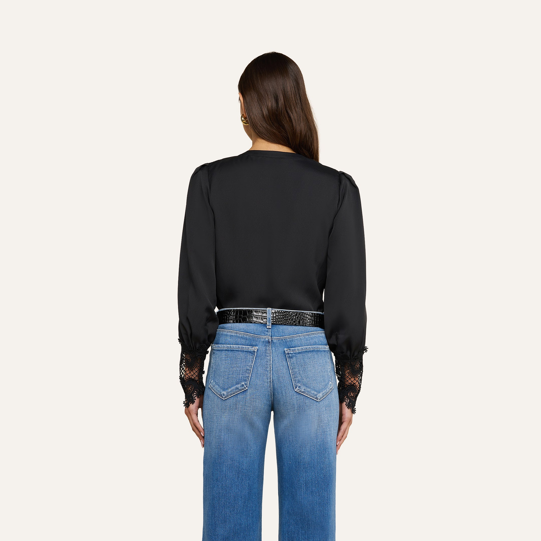 Una donna è di spalle e indossa la camicetta Ava — un top nero di ispirazione vintage con polsini in pizzo guipure smerlato — abbinata a jeans blu a vita alta e una cintura nera. I suoi capelli castani lisci ricadono sciolti su uno sfondo beige a tinta unita.