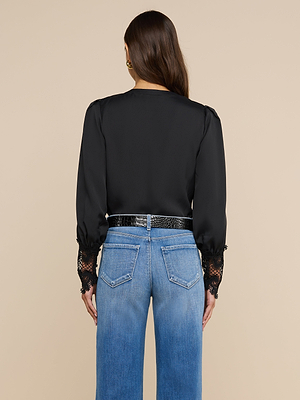Una donna è di spalle e indossa la camicetta Ava — un top nero di ispirazione vintage con polsini in pizzo guipure smerlato — abbinata a jeans blu a vita alta e una cintura nera. I suoi capelli castani lisci ricadono sciolti su uno sfondo beige a tinta unita.