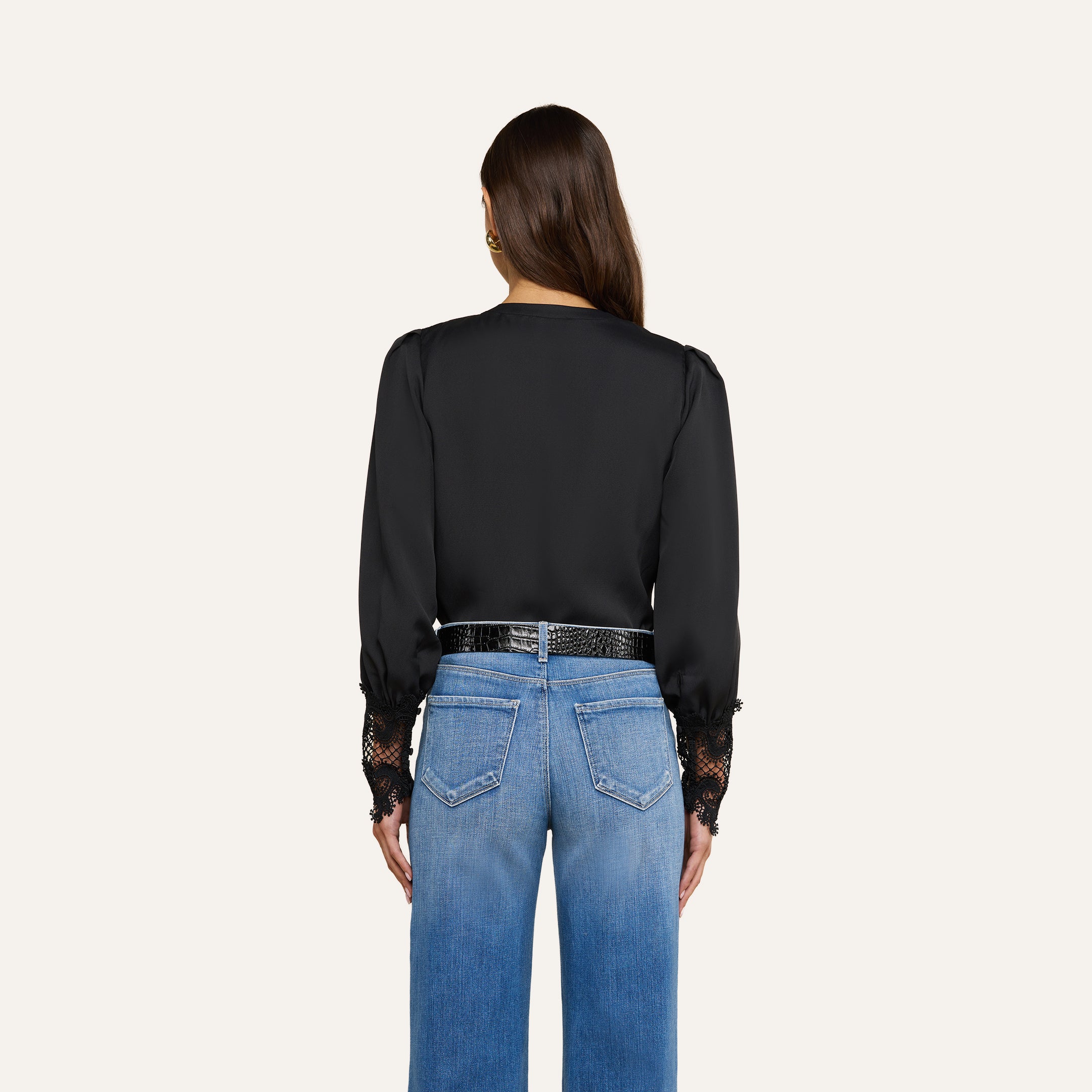 Una donna è di spalle e indossa la camicetta Ava — un top nero di ispirazione vintage con polsini in pizzo guipure smerlato — abbinata a jeans blu a vita alta e una cintura nera. I suoi capelli castani lisci ricadono sciolti su uno sfondo beige a tinta unita.