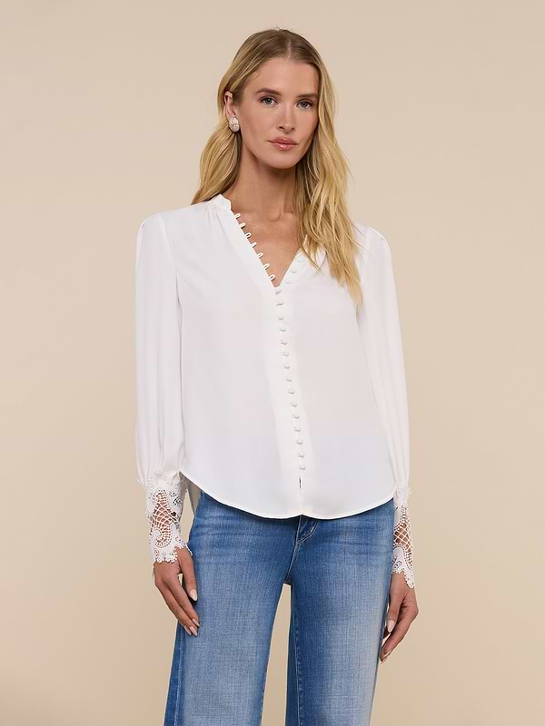 A woman wearing the Ava Blouse—an ivory, vintage-inspired top with scalloped guipure lace cuffs—and blue jeans stands against a plain light background, facing forward with a neutral expression.