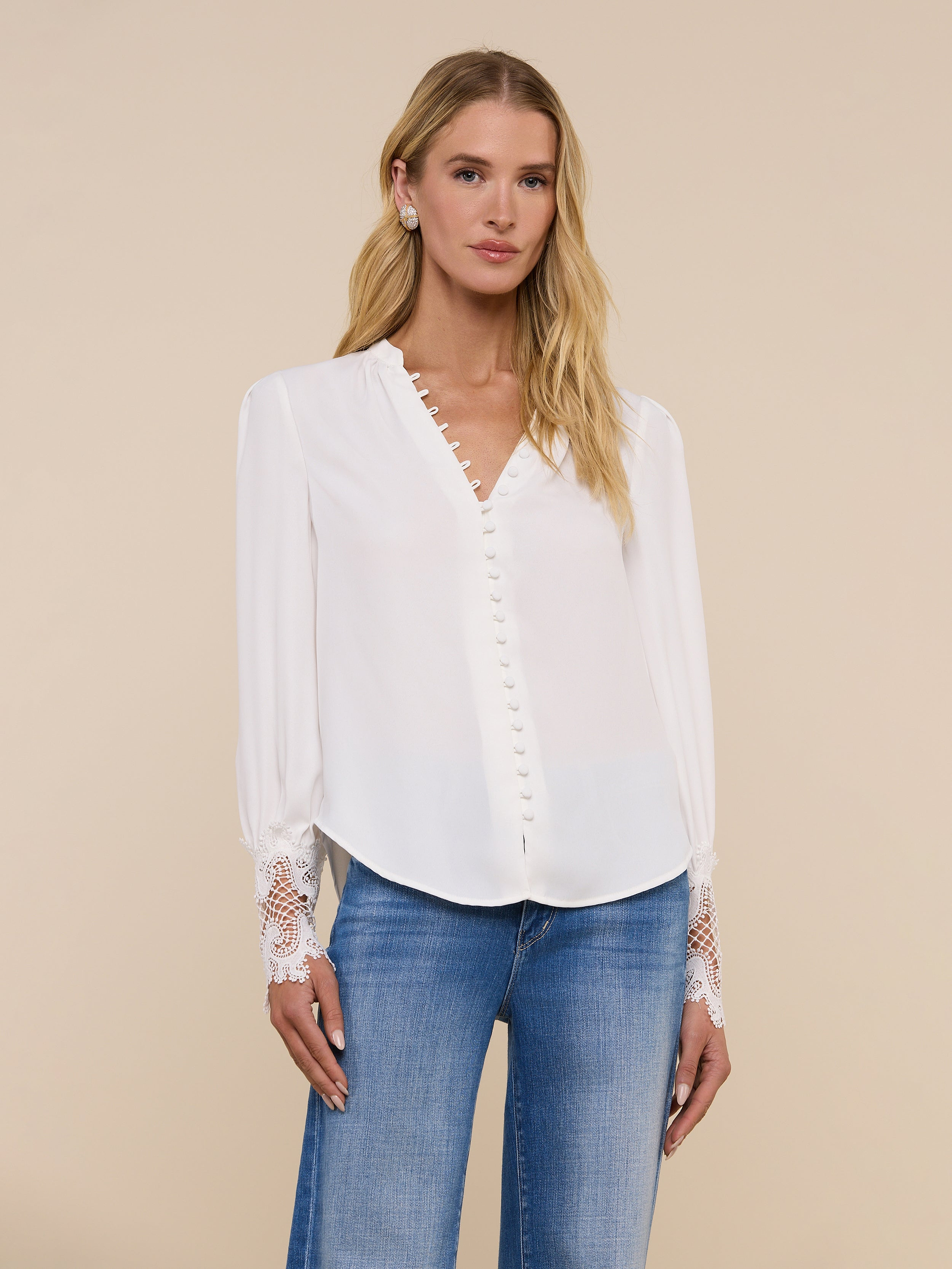 A woman wearing the Ava Blouse—an ivory, vintage-inspired top with scalloped guipure lace cuffs—and blue jeans stands against a plain light background, facing forward with a neutral expression.