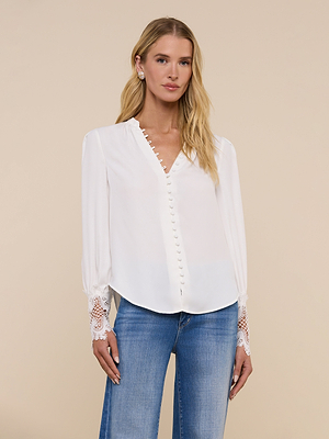 A woman with long blonde hair wears the Ava Blouse, a vintage-inspired ivory top with scalloped guipure lace cuffs, paired with blue jeans against a plain beige background.