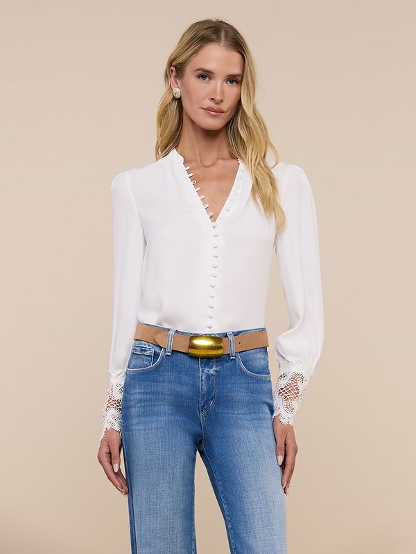 A woman with long blonde hair wears the Ava Blouse, a vintage-inspired ivory top featuring scalloped guipure lace cuffs, paired with blue jeans. She stands facing forward against a plain light background.