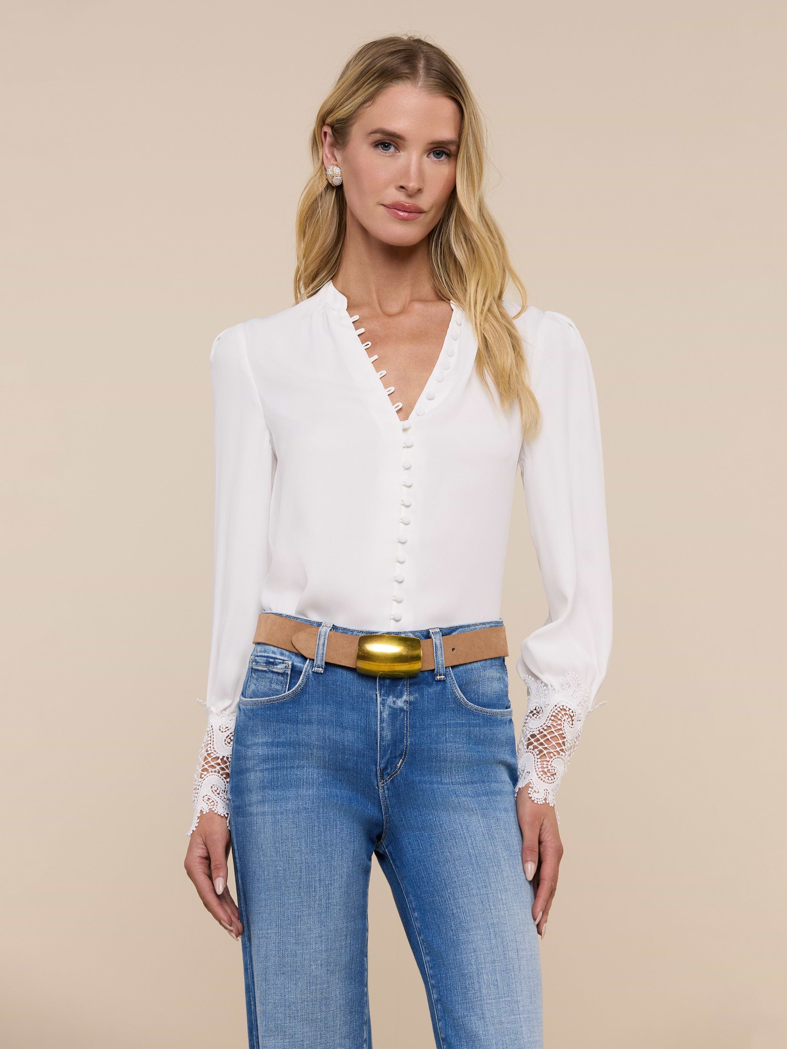 A woman with long blonde hair wears the Ava Blouse, a vintage-inspired ivory top featuring scalloped guipure lace cuffs, paired with blue jeans. She stands facing forward against a plain light background.