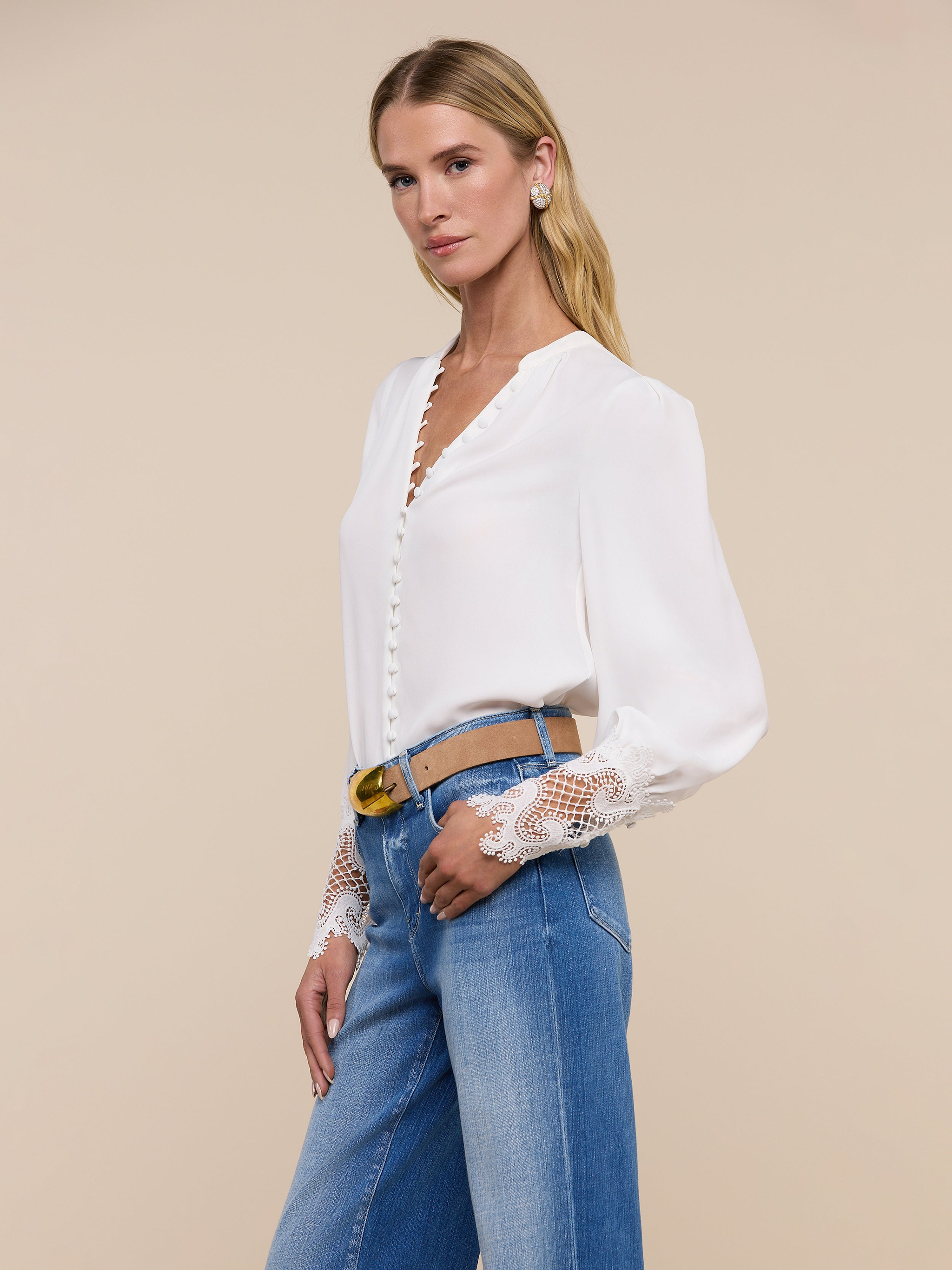 A woman poses against a plain, light background wearing the Ava Blouse—an ivory, vintage-inspired top featuring scalloped guipure lace cuffs—paired with blue high-waisted jeans.