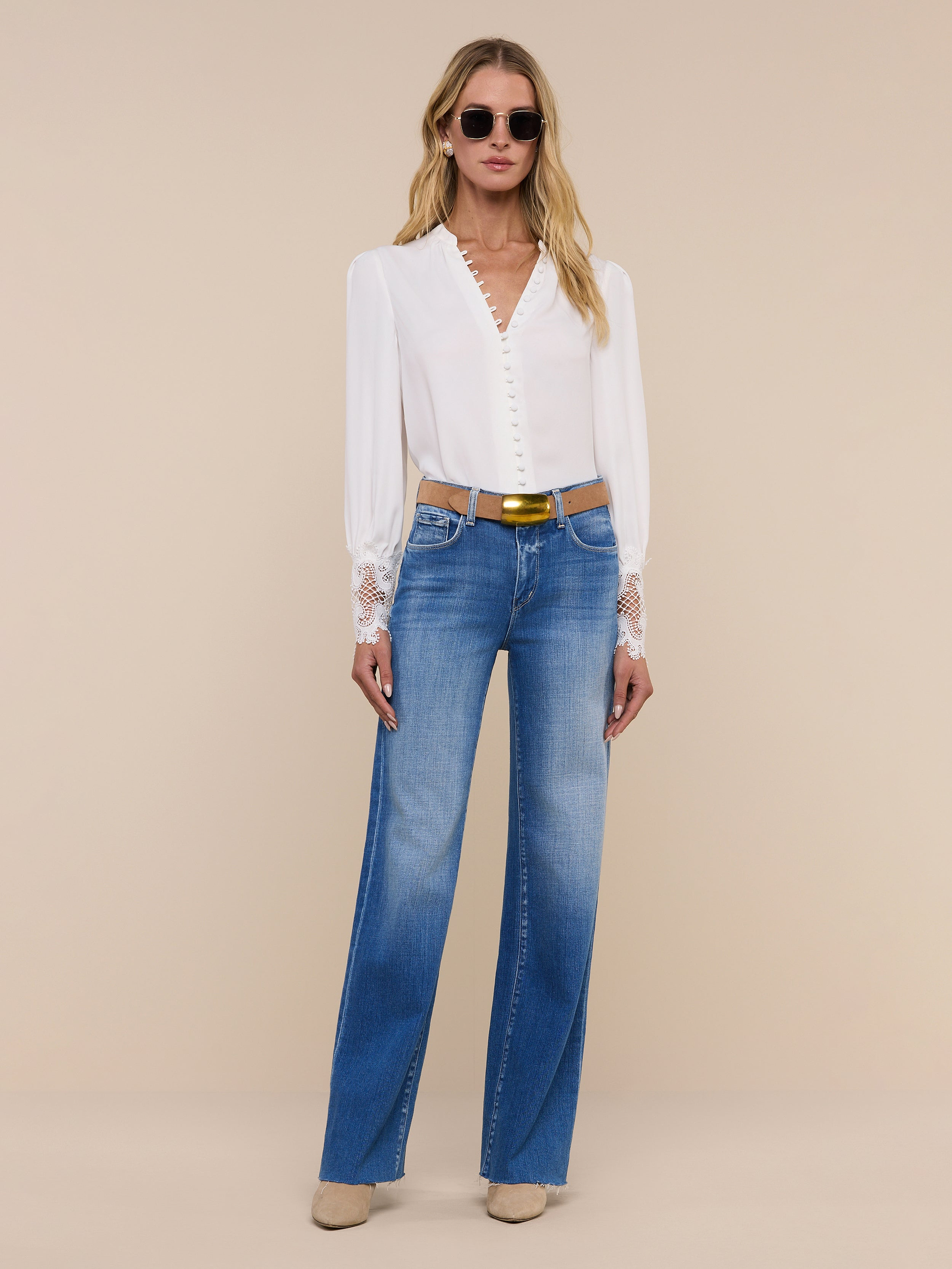 A woman wearing sunglasses, the Ava Blouse—a vintage-inspired ivory top with scalloped guipure lace cuffs—blue high-waisted jeans, a black belt, and tan shoes stands confidently against a plain white background.