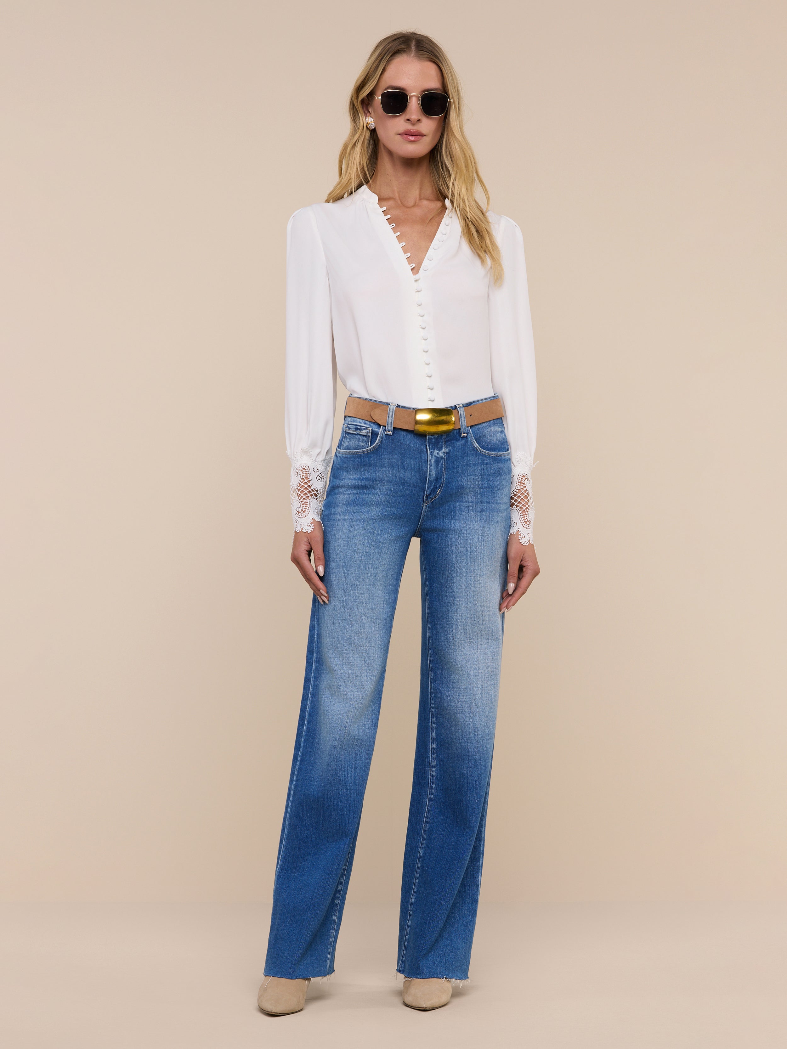 A woman wearing sunglasses, the Ava Blouse—a vintage-inspired ivory top with scalloped guipure lace cuffs—blue high-waisted jeans, a black belt, and tan shoes stands confidently against a plain white background.