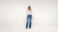 Woman stands facing forward against a plain white background, wearing sunglasses, a white long-sleeve blouse, blue wide-leg jeans, a yellow belt, and beige shoes.
