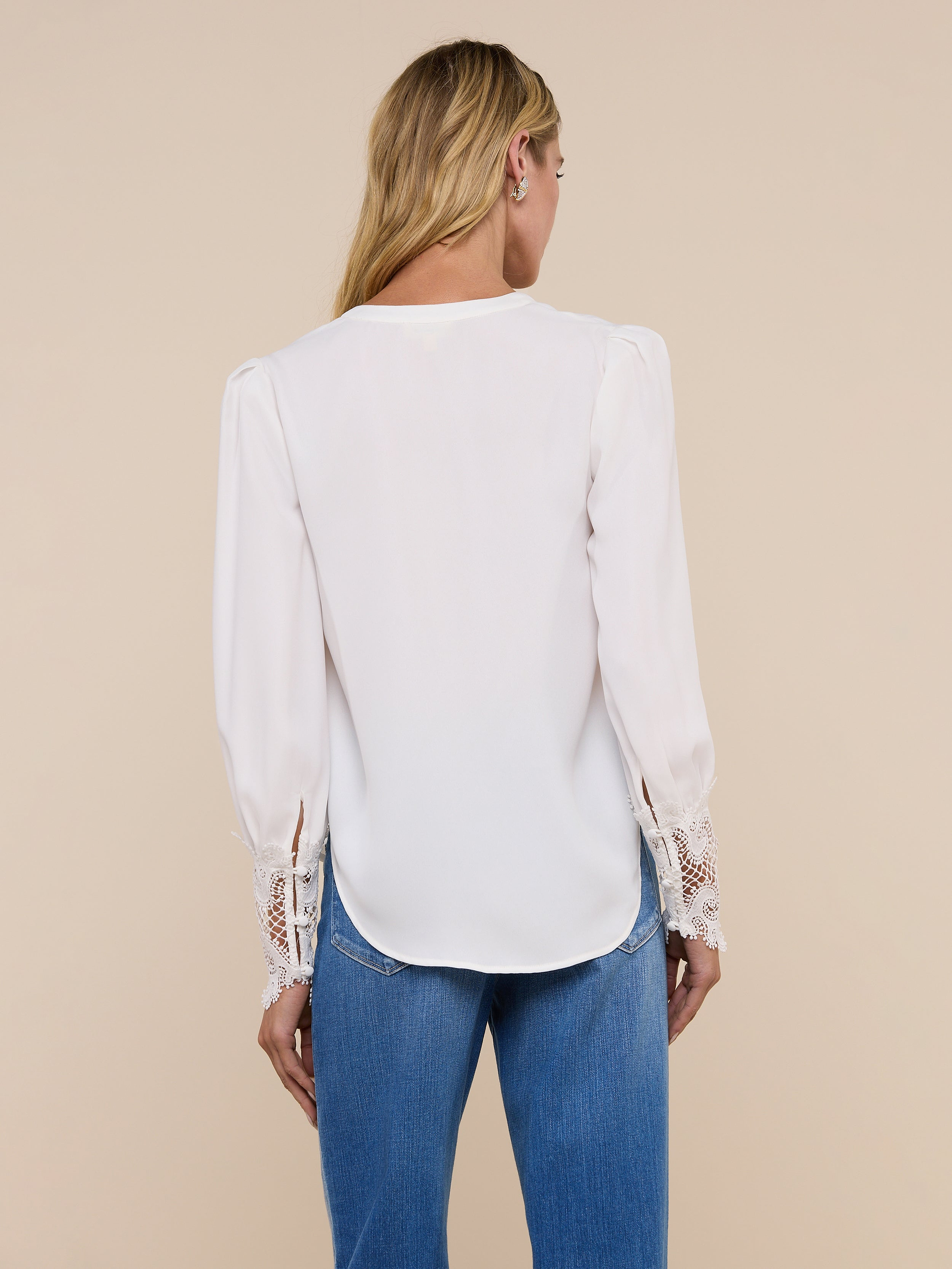 A woman with long blonde hair is seen from behind, wearing the vintage-inspired Ava Blouse in ivory with scalloped guipure lace cuffs, paired with blue jeans, against a plain light background.