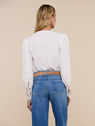 A woman stands facing away, wearing the Ava Blouse—a vintage-inspired ivory top featuring scalloped guipure lace cuffs—paired with blue jeans. Her blonde hair is worn down, and the background is plain and light-colored.
