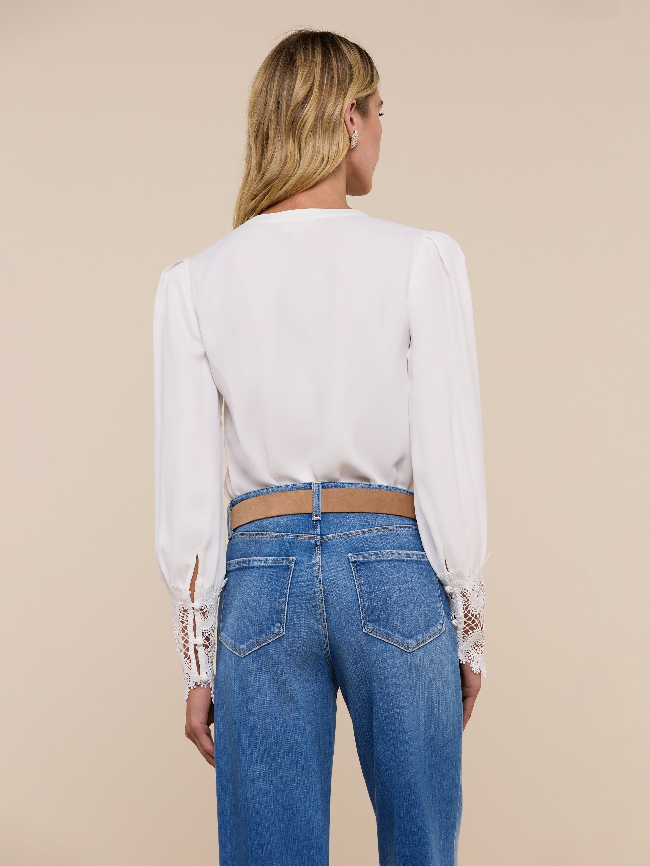 A woman stands facing away, wearing the Ava Blouse—a vintage-inspired ivory top featuring scalloped guipure lace cuffs—paired with blue jeans. Her blonde hair is worn down, and the background is plain and light-colored.