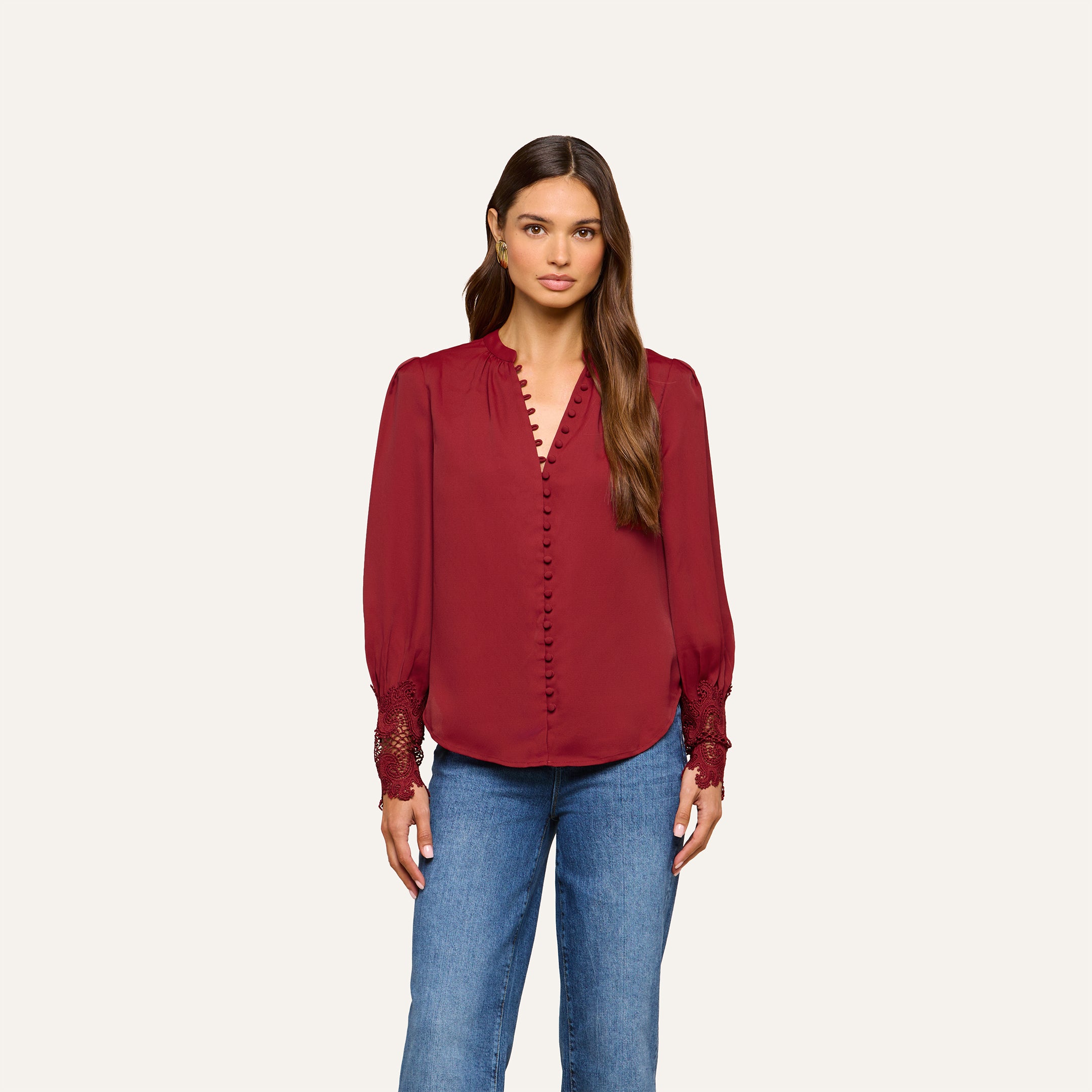 A woman stands against a plain background, wearing the Ava Blouse—a vintage-inspired top in deep red featuring puff sleeves, button details, and scalloped lace cuffs—paired with blue jeans.