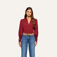 A woman wears the Ava Blouse, a vintage-inspired long-sleeve burgundy top featuring button details and embroidered cuffs, styled with blue jeans and a tan belt, standing against a plain light background.