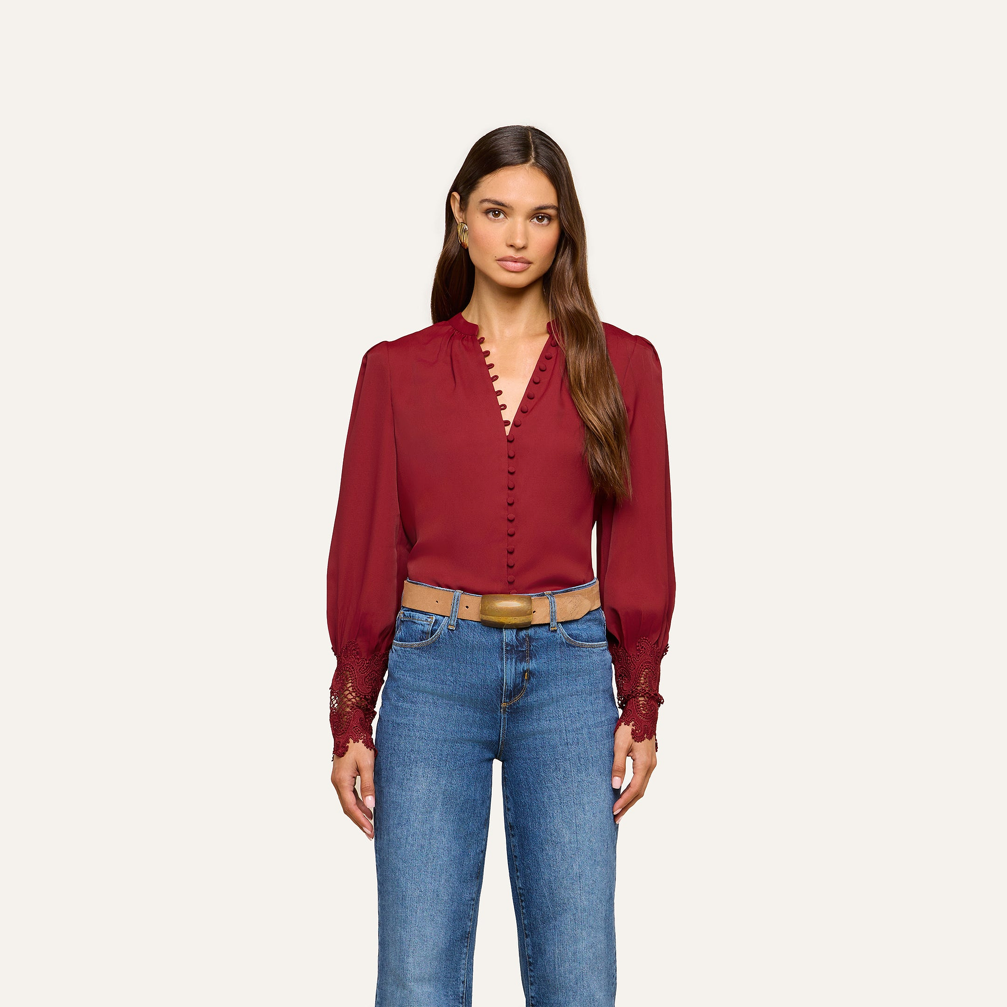 A woman wears the Ava Blouse, a vintage-inspired long-sleeve burgundy top featuring button details and embroidered cuffs, styled with blue jeans and a tan belt, standing against a plain light background.