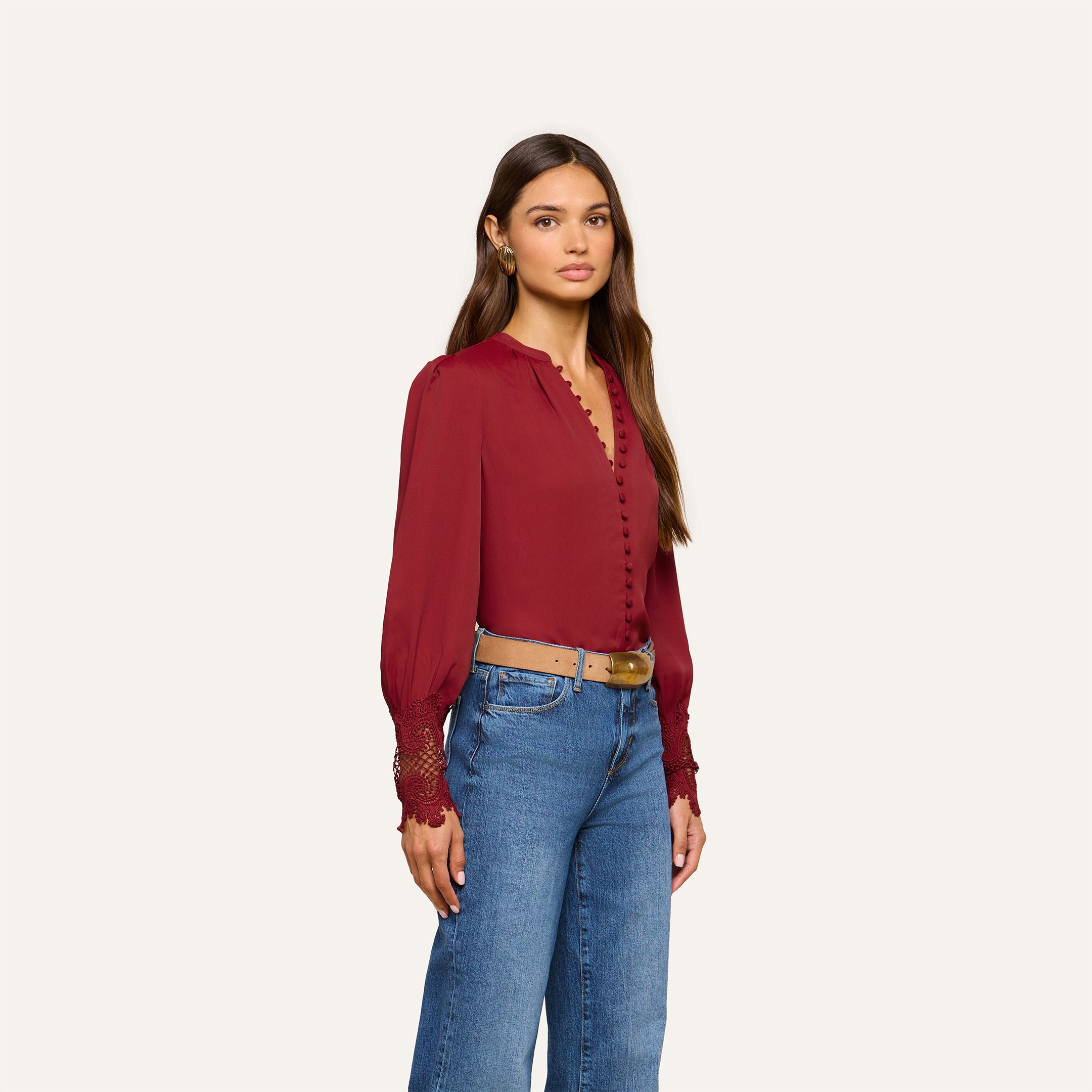 A woman with long brown hair wears the Ava Blouse, a vintage-inspired maroon top featuring scalloped lace cuffs and puff sleeves, paired with blue jeans, standing against a plain light background.