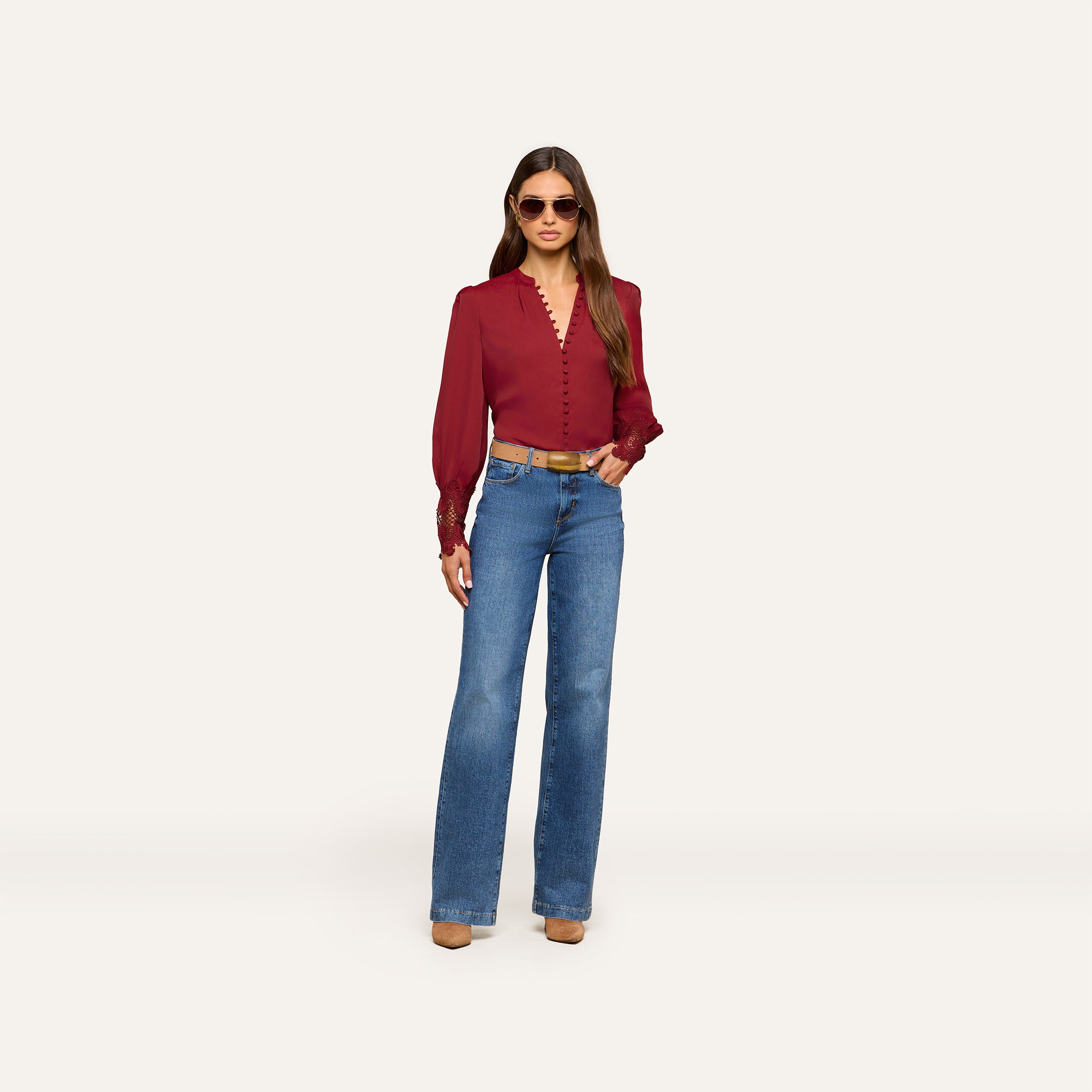 A person wearing the Ava Blouse—a vintage-inspired burgundy top with puff sleeves—paired with blue high-waisted jeans, brown heeled shoes, and sunglasses stands against a plain light background.