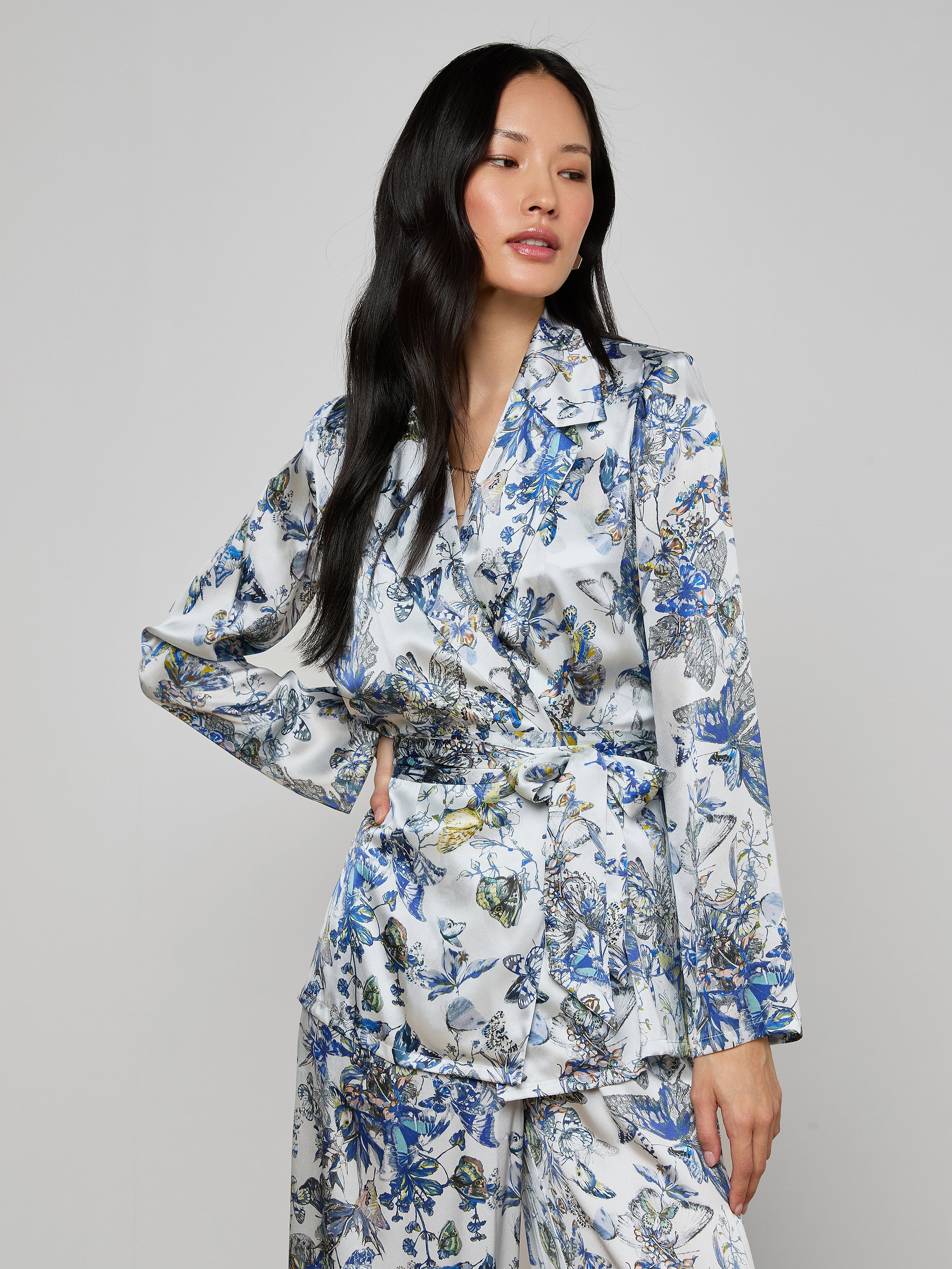 A woman with long black hair wears the Ciara Silk Robe Top in a blue, green, and white floral pattern, standing against a plain light gray background.