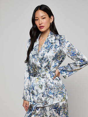 A woman with long black hair poses against a plain light background, wearing the Ciara Silk Robe Top—a white belted wrap-style blazer—paired with matching blue and yellow floral pants, one hand on her hip.
