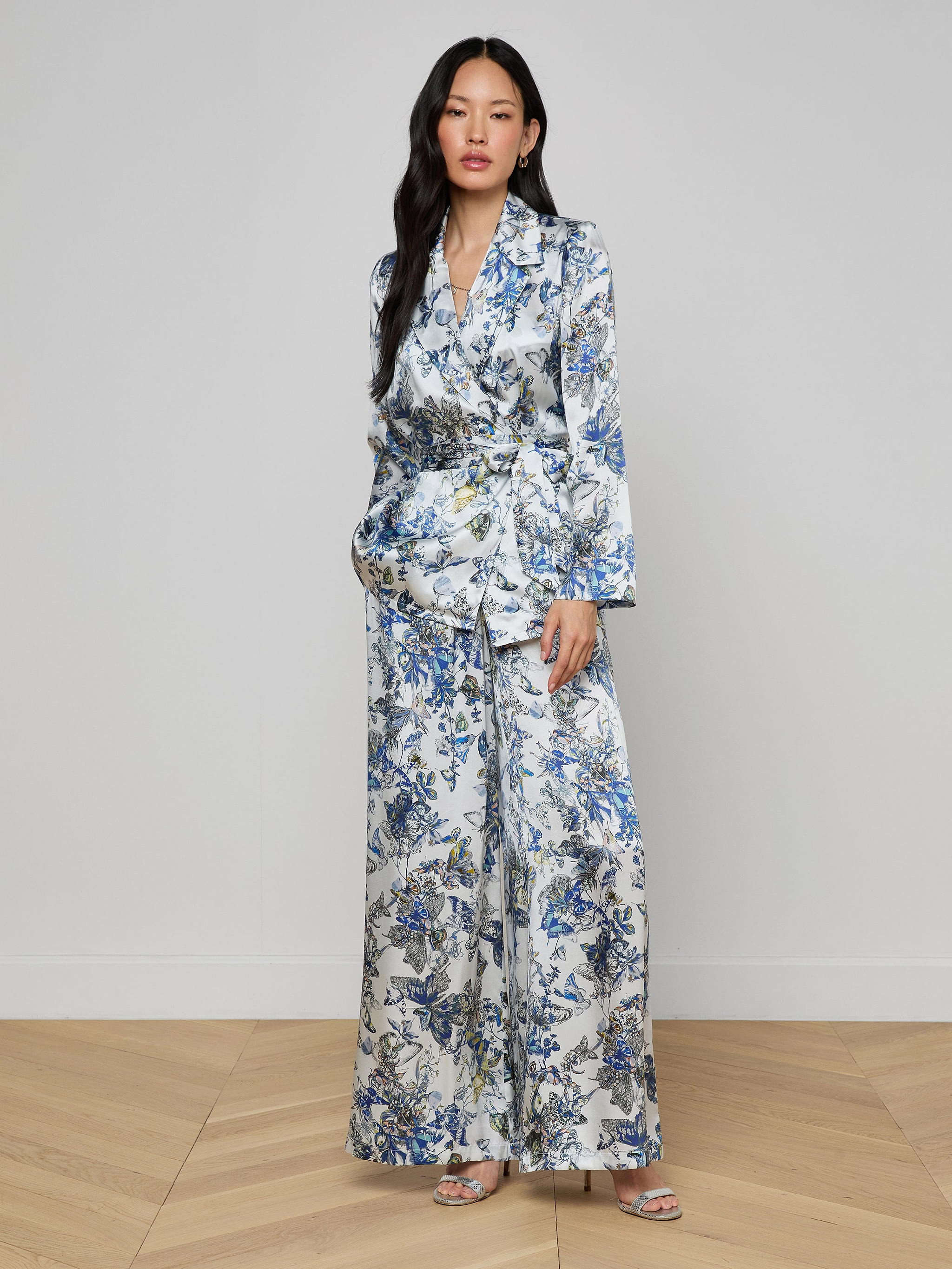 A woman with long dark hair stands against a light wall, hands in her pockets, wearing the Ciara Silk Robe Top—an elegant silk charmeuse piece with a blue, white, and yellow floral pattern—and open-toe heels.