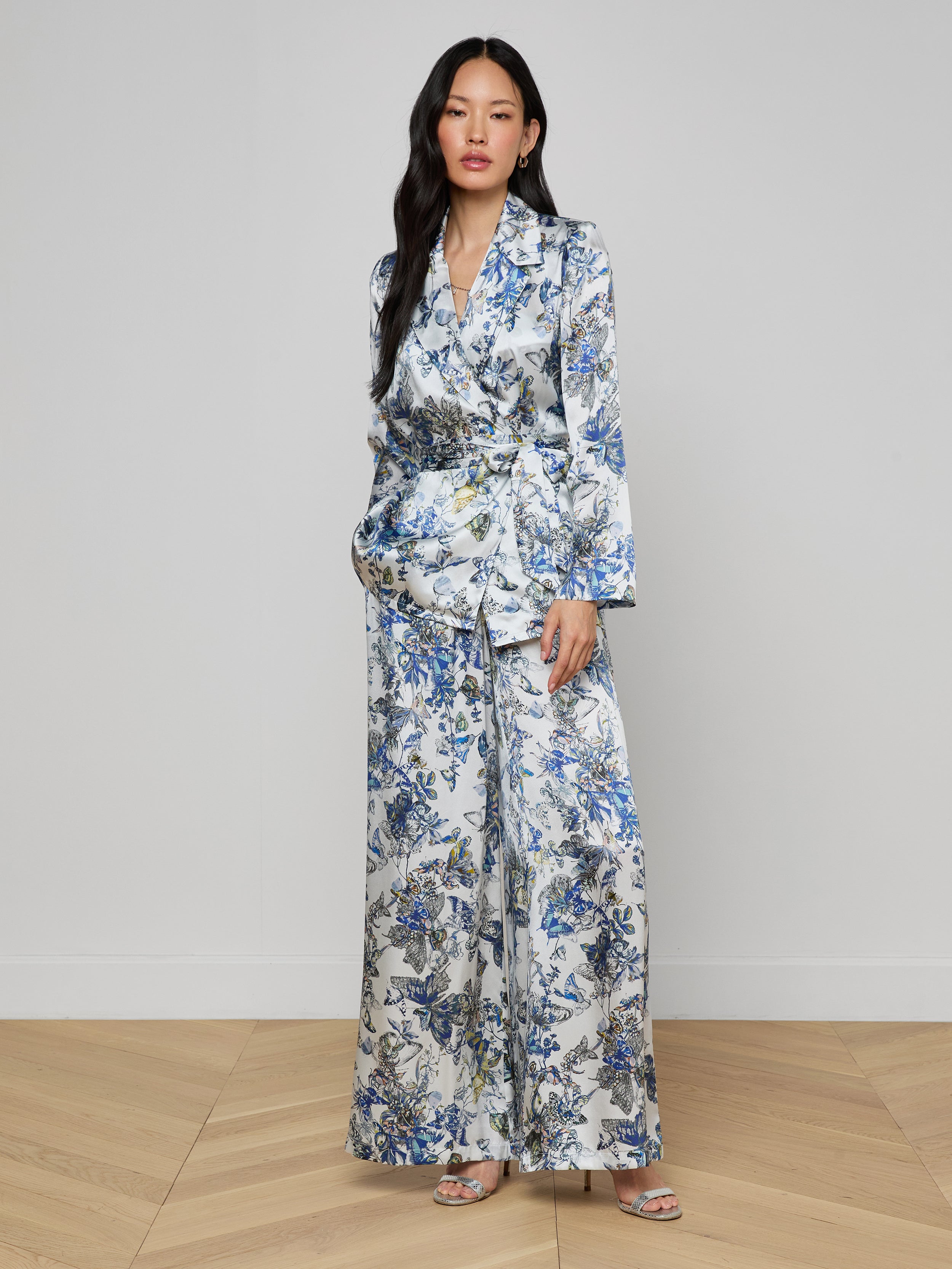 A woman with long dark hair stands against a light wall, hands in her pockets, wearing the Ciara Silk Robe Top—an elegant silk charmeuse piece with a blue, white, and yellow floral pattern—and open-toe heels.