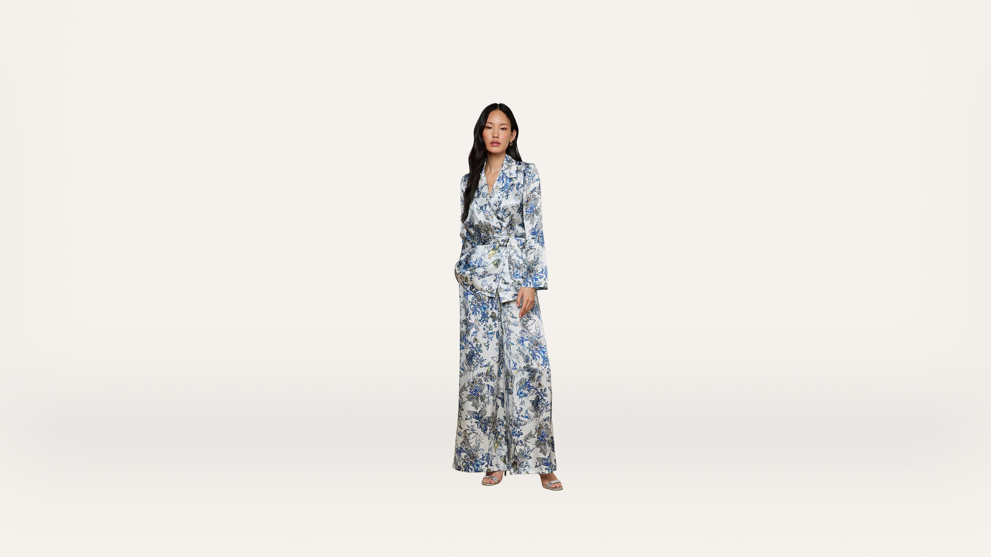 A woman stands against a plain white background, wearing a blue and white floral patterned suit with wide-legged pants and sandals.