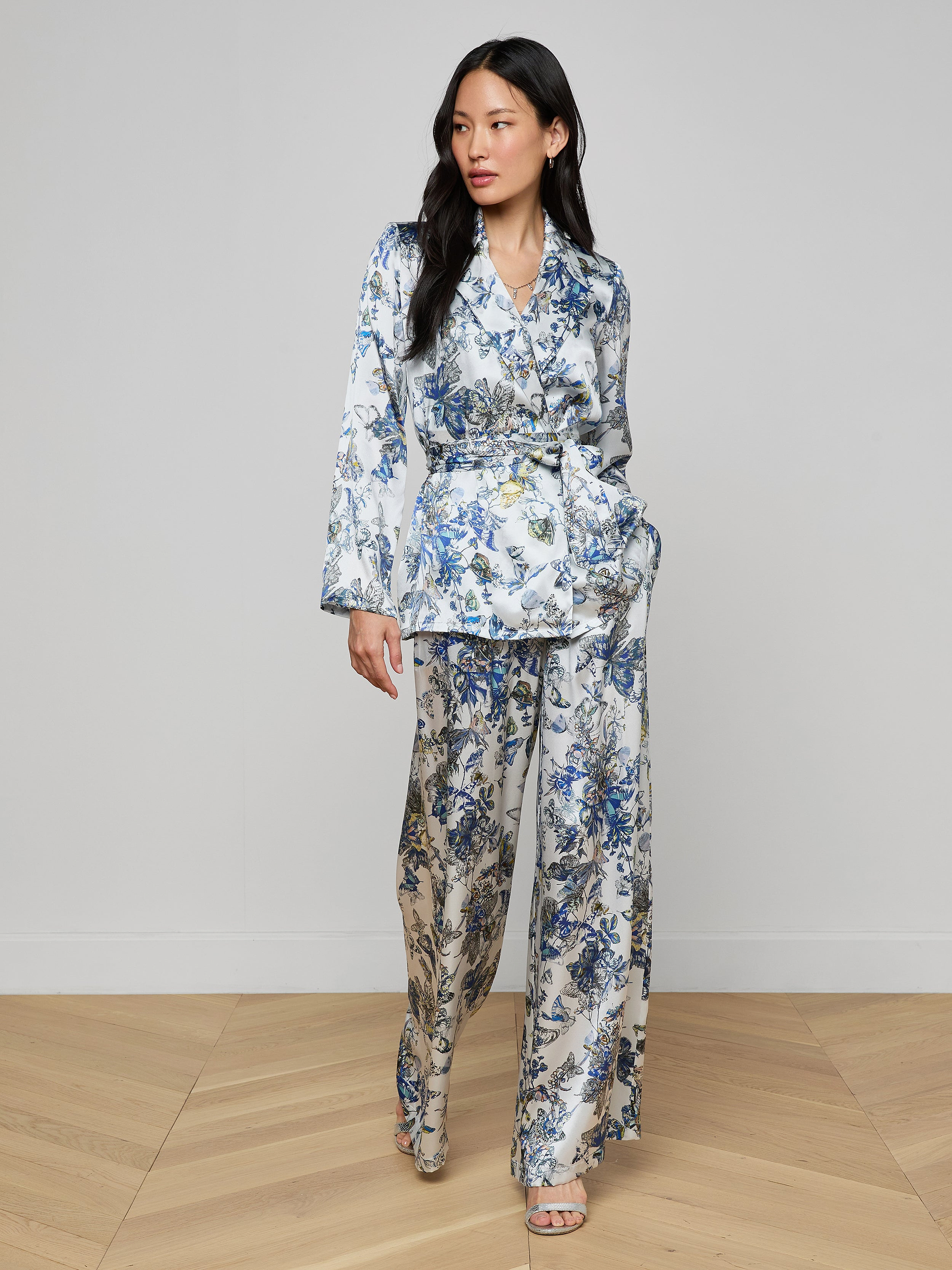 A woman with long dark hair poses confidently on a wooden floor in the Ciara Silk Robe Top, styled as part of a matching white pantsuit with blue floral prints and a belted waist, paired with open-toe heels against a plain light background.