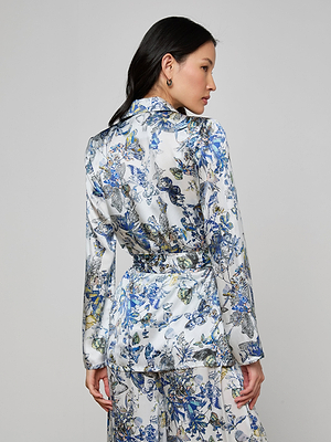 A woman with long dark hair wears the Ciara Silk Robe Top, white with a blue and gray floral pattern. She faces away, looking to the side against a plain light gray background.