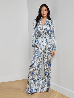 A woman with long dark hair poses confidently indoors against a white wall, wearing the Ciara Silk Robe Top—a silky silk charmeuse top with a blue, white, and yellow floral pattern. She has one hand in her pocket.
