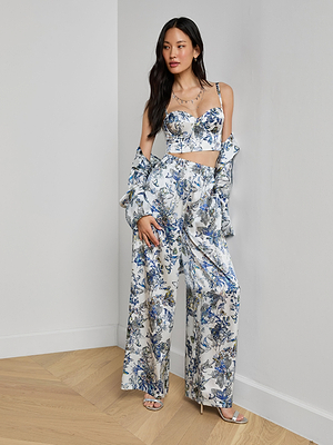A woman stands against a white wall, wearing the Ciara Silk Robe Top—a floral print silk charmeuse crop top—paired with matching wide-leg pants, an open shirt in white and blue tones, heeled sandals, and layered necklaces.