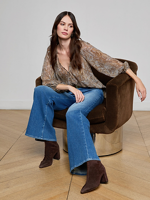 A woman with long brown hair sits in a brown swivel chair, wearing a paisley blouse, blue flared jeans, and brown heeled boots, with a neutral expression in a modern room with wooden floors.