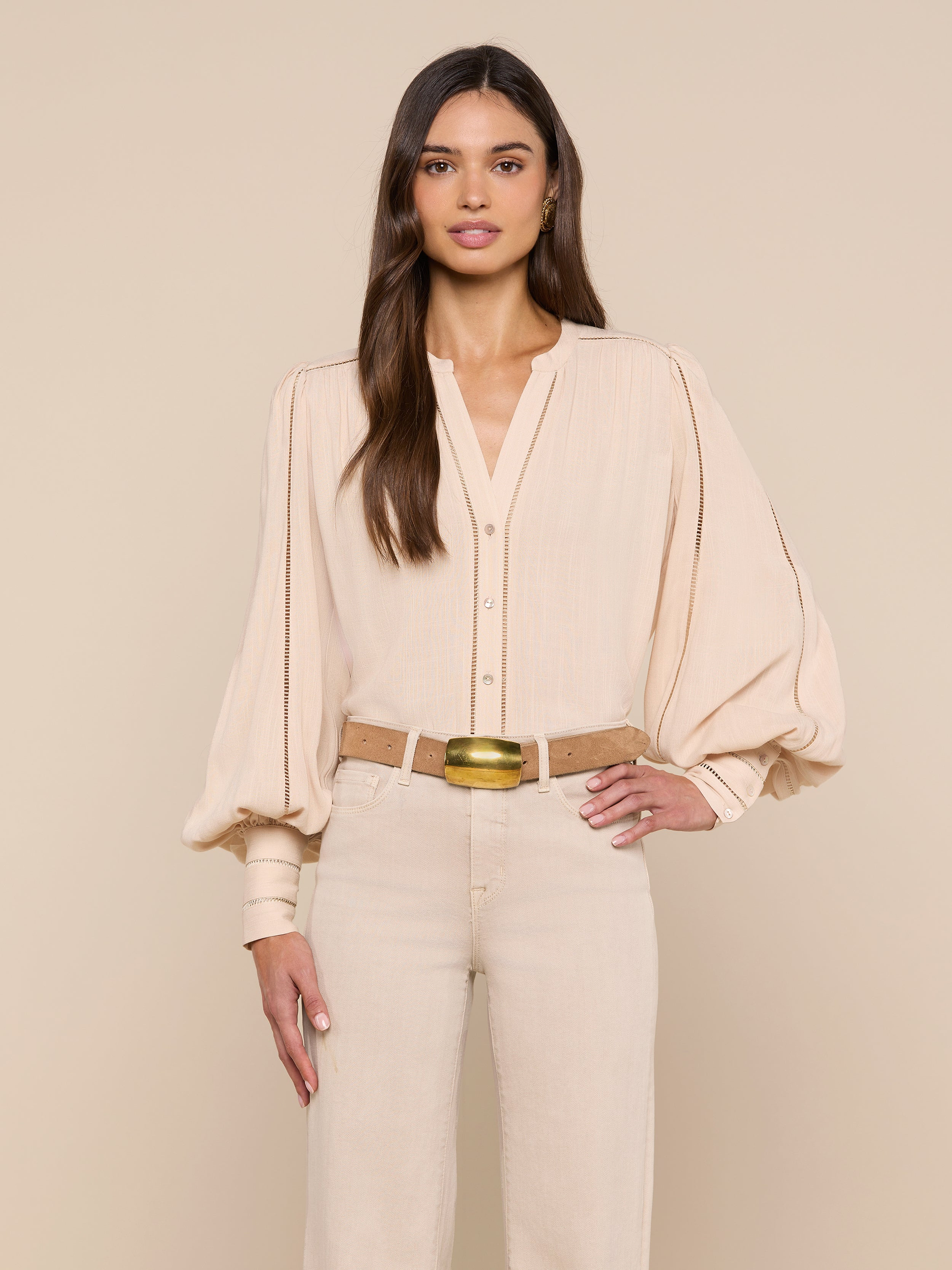 A woman with long brown hair stands facing forward, wearing the beige Kiera Blouse with blouson sleeves and beige high-waisted pants. She has one hand on her hip against a plain white background.