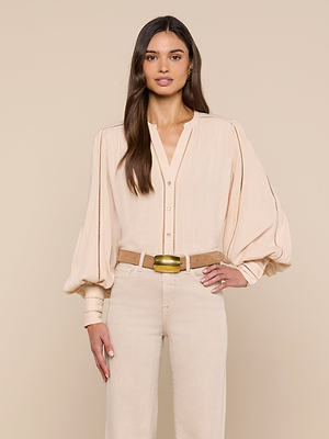A woman with long brown hair and a neutral expression stands against a beige background, wearing the Kiera Blouse—a light beige bohemian top with blouson sleeves—paired with beige high-waisted pants and a tan belt with a gold buckle.
