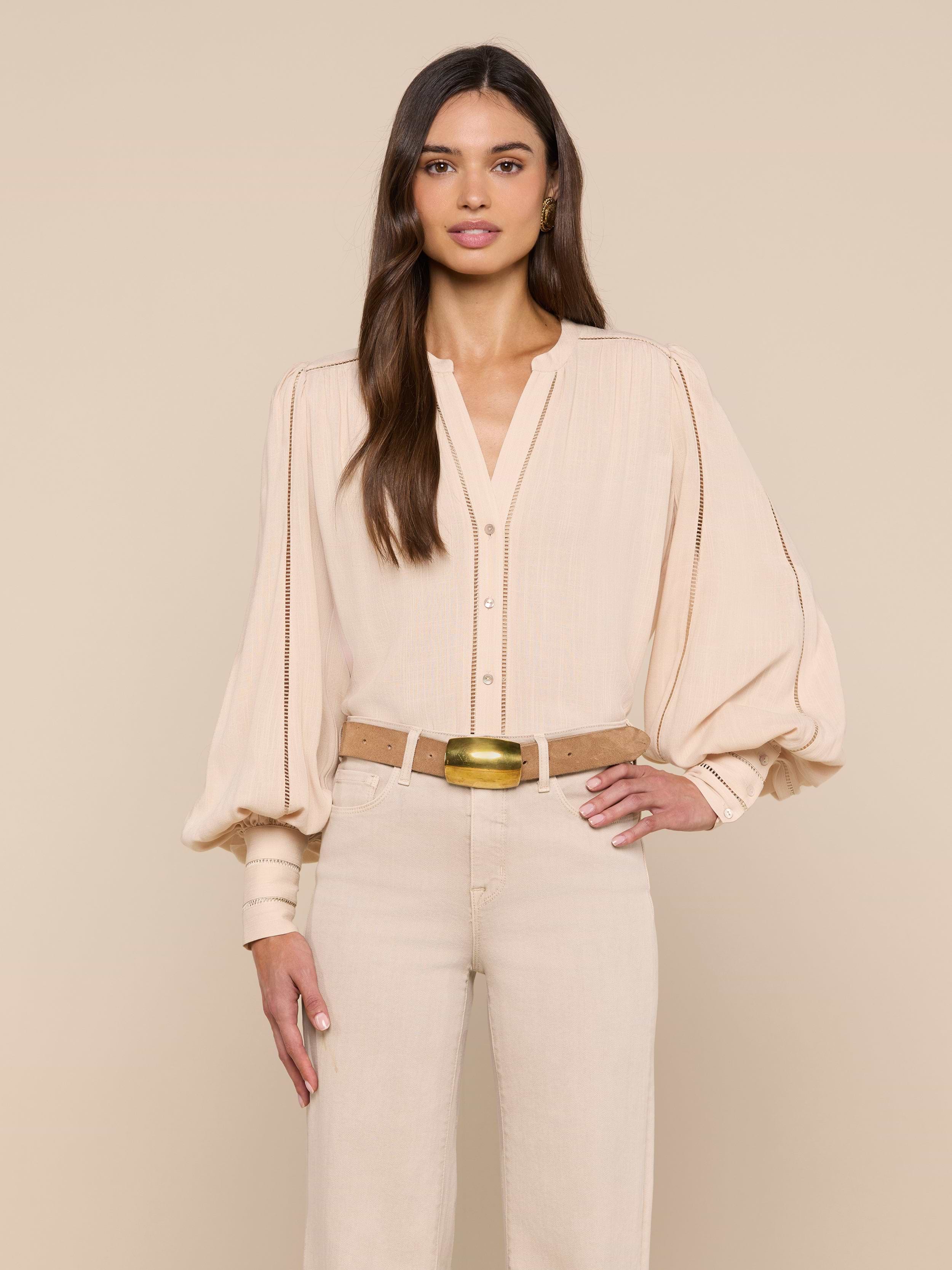 A woman with long brown hair stands facing forward, wearing the beige Kiera Blouse with blouson sleeves and beige high-waisted pants. She has one hand on her hip against a plain white background.