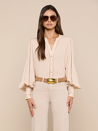 A woman wearing sunglasses, the Kiera Blouse—a beige bohemian top with blouson sleeves—beige pants, and a tan belt stands facing forward against a plain light background.