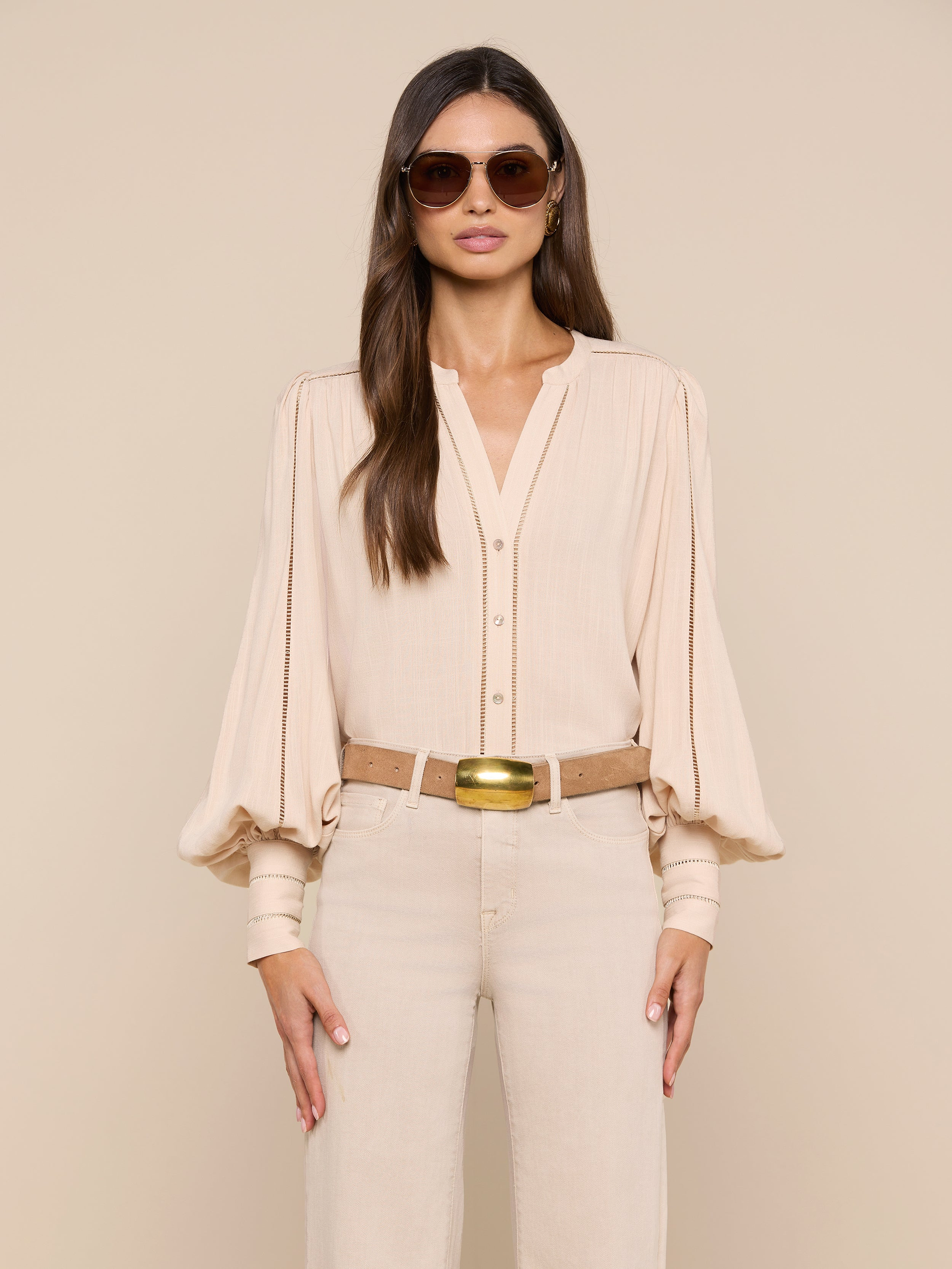 A woman wearing sunglasses, the Kiera Blouse—a beige bohemian top with blouson sleeves—beige pants, and a tan belt stands facing forward against a plain light background.