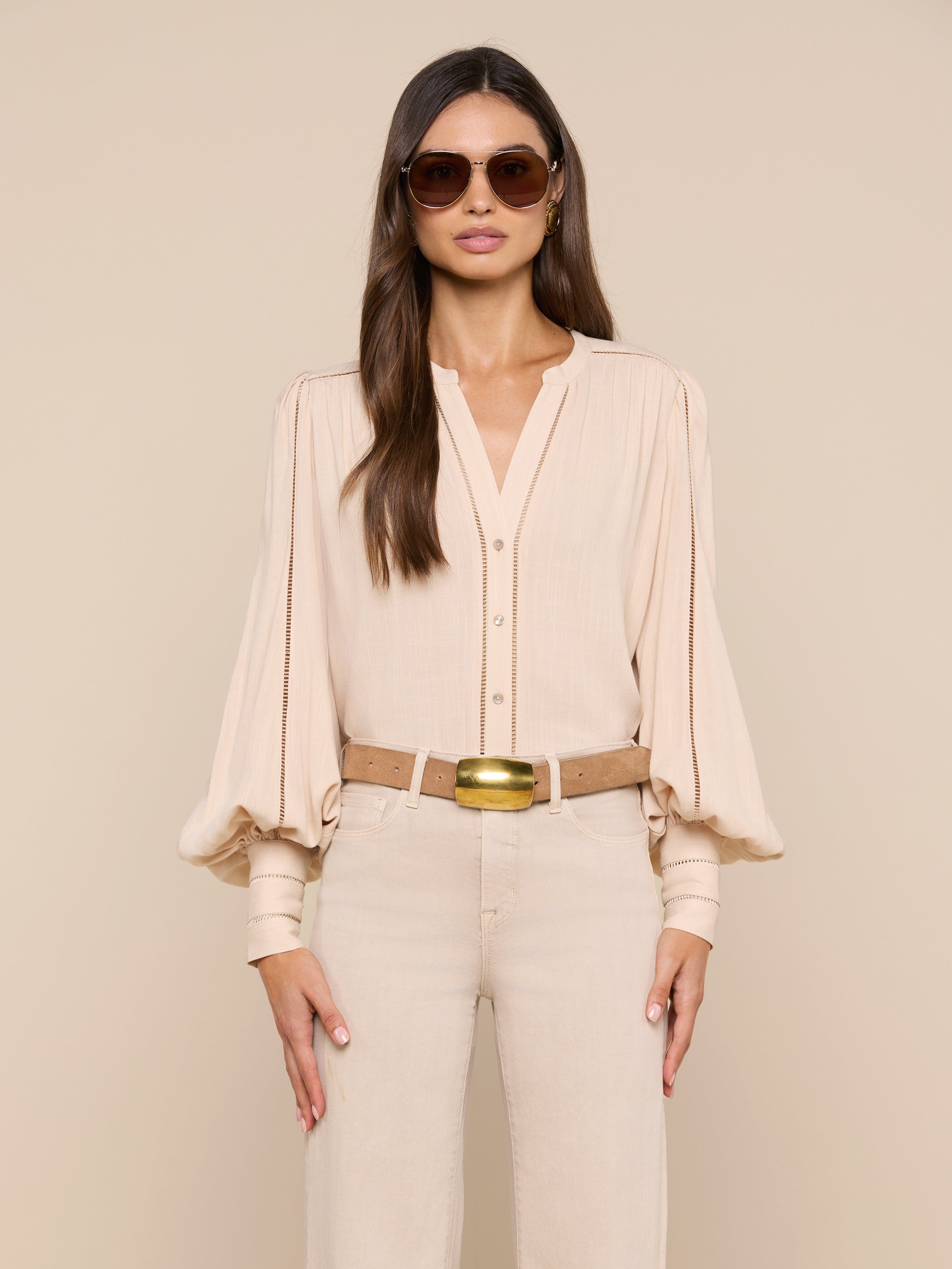 A woman wearing sunglasses, the Kiera Blouse—a beige bohemian top with blouson sleeves—beige pants, and a tan belt stands facing forward against a plain light background.