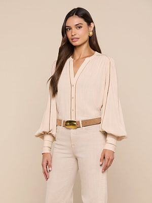 A woman with long brown hair wears the Kiera Blouse—a beige bohemian top with blouson sleeves—paired with beige pants, a tan belt with a gold buckle, and gold hoop earrings, standing against a beige background.