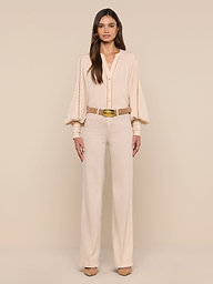 A woman with long, straight brown hair and a neutral expression stands against a plain white background wearing the Kiera Blouse—a beige, bohemian-style top with blouson sleeves—paired with high-waisted beige trousers, a gold belt, and beige heels.