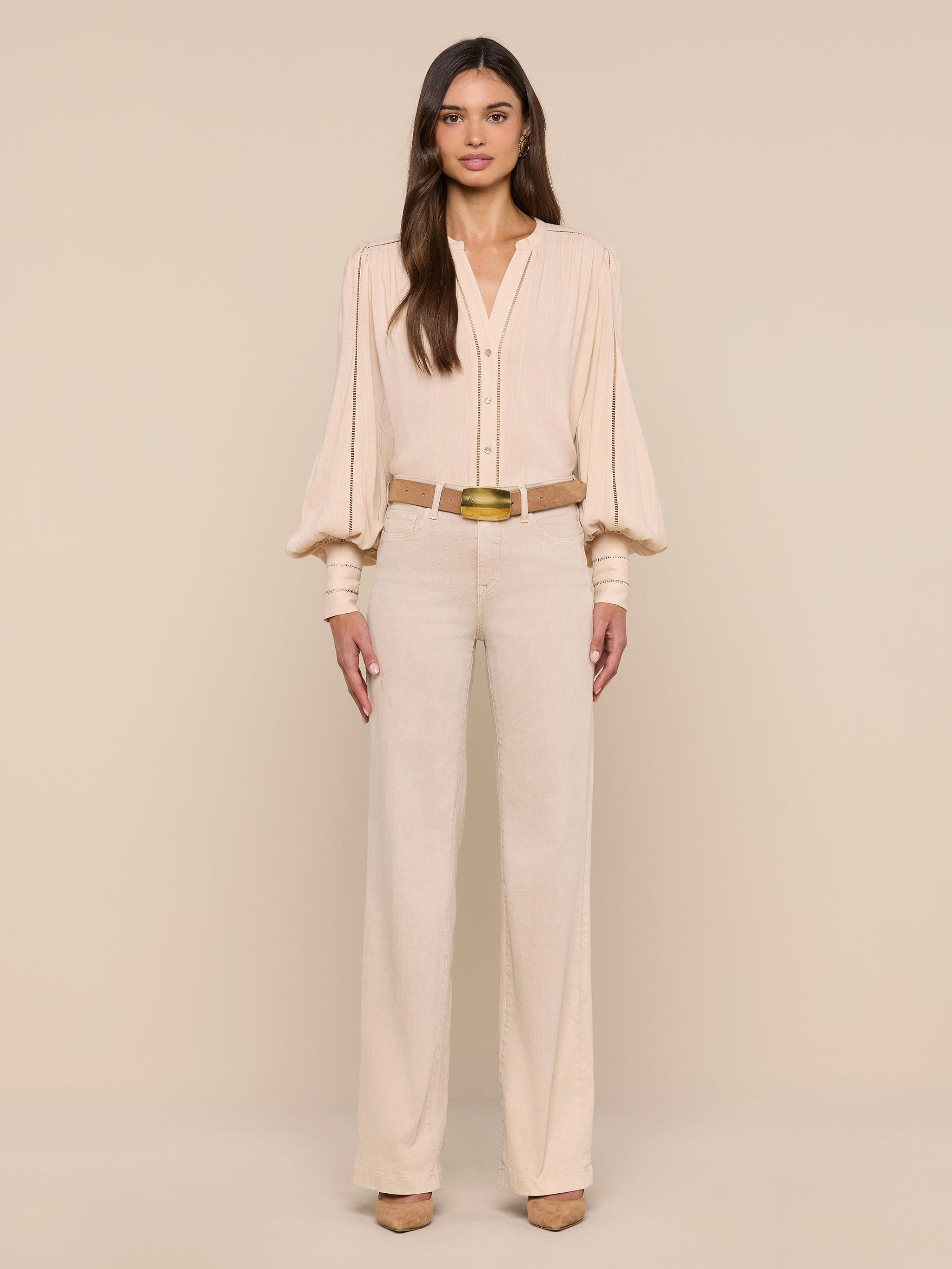 A woman with long, straight brown hair and a neutral expression stands against a plain white background wearing the Kiera Blouse—a beige, bohemian-style top with blouson sleeves—paired with high-waisted beige trousers, a gold belt, and beige heels.