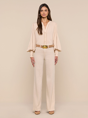 A woman faces forward against a beige background, wearing the light beige Kiera Blouse with long blouson sleeves, wide-leg beige pants, a tan belt with a large buckle, and beige platform shoes.