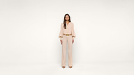 A woman stands against a plain white background, wearing a light beige blouse with long sleeves, beige pants, matching heels, and a brown belt. She faces forward with her arms relaxed at her sides.