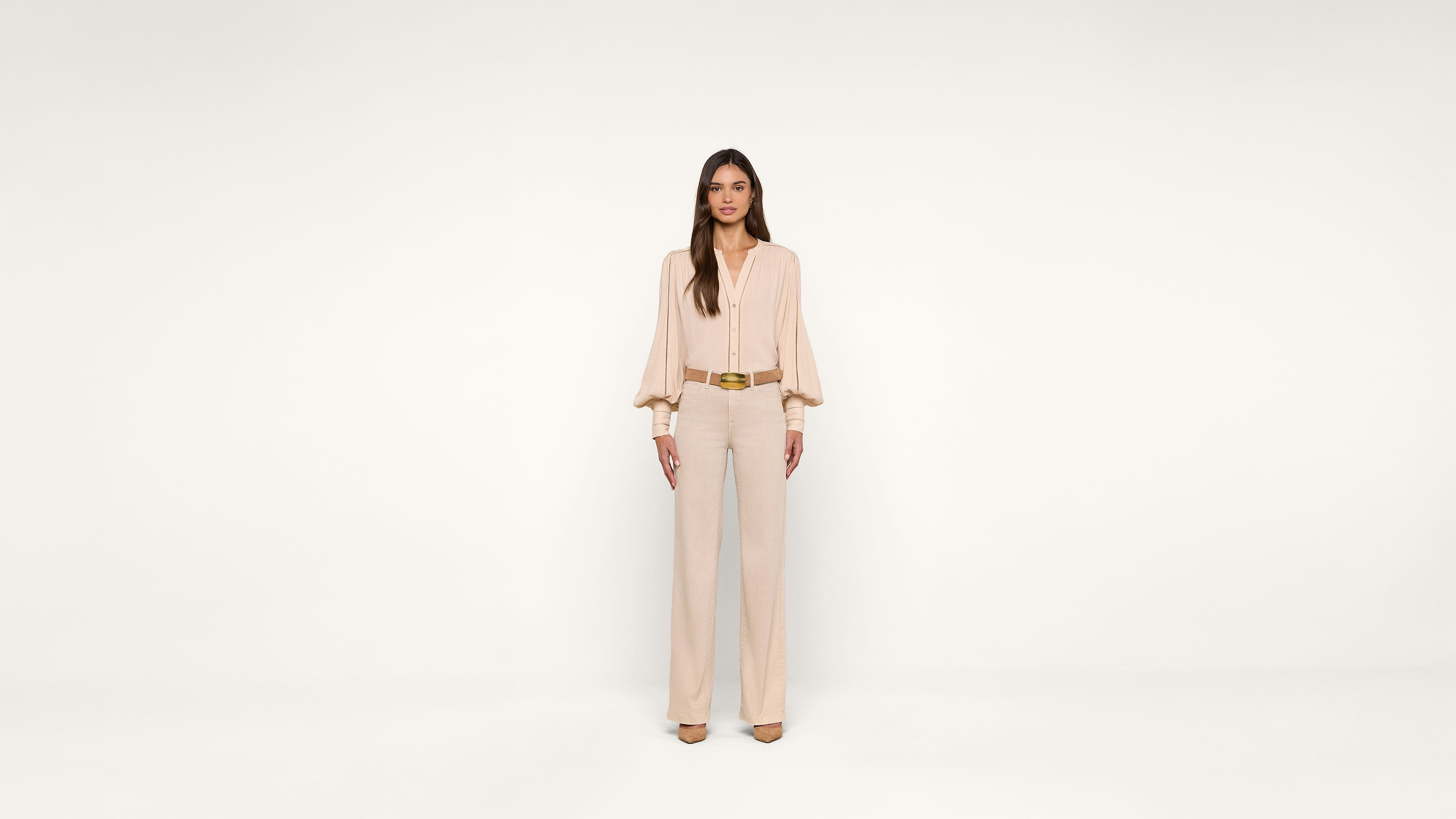 A woman stands against a plain white background, wearing a light beige blouse with long sleeves, beige pants, matching heels, and a brown belt. She faces forward with her arms relaxed at her sides.