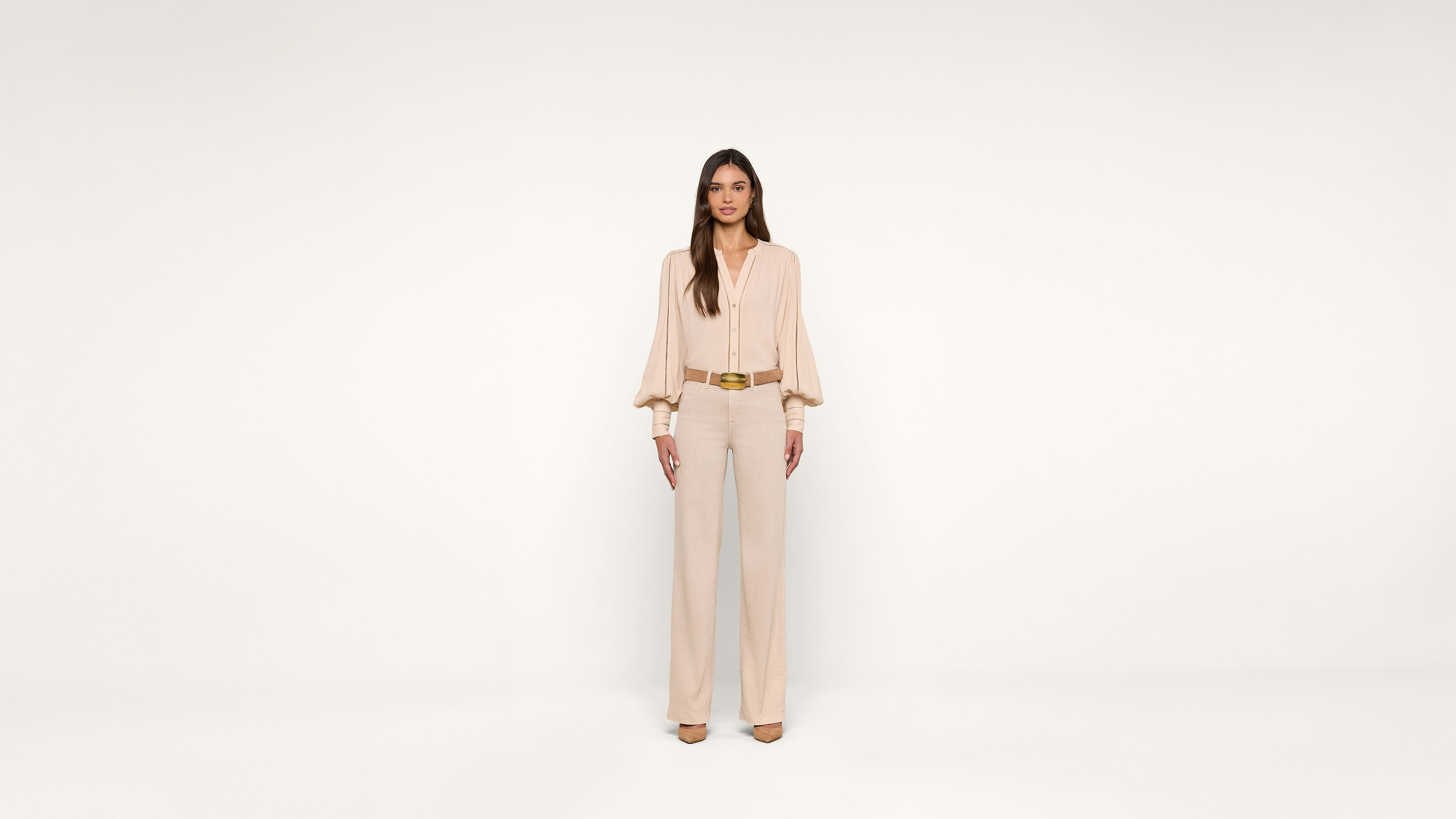 A woman stands against a plain white background, wearing a light beige blouse with long sleeves, beige pants, matching heels, and a brown belt. She faces forward with her arms relaxed at her sides.