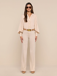 A woman stands confidently in a neutral studio, wearing the Kiera Blouse—a beige bohemian top with blouson sleeves—paired with matching high-waisted trousers, a brown belt, beige heels, and large sunglasses. Her long straight hair and poised stance complete the look.
