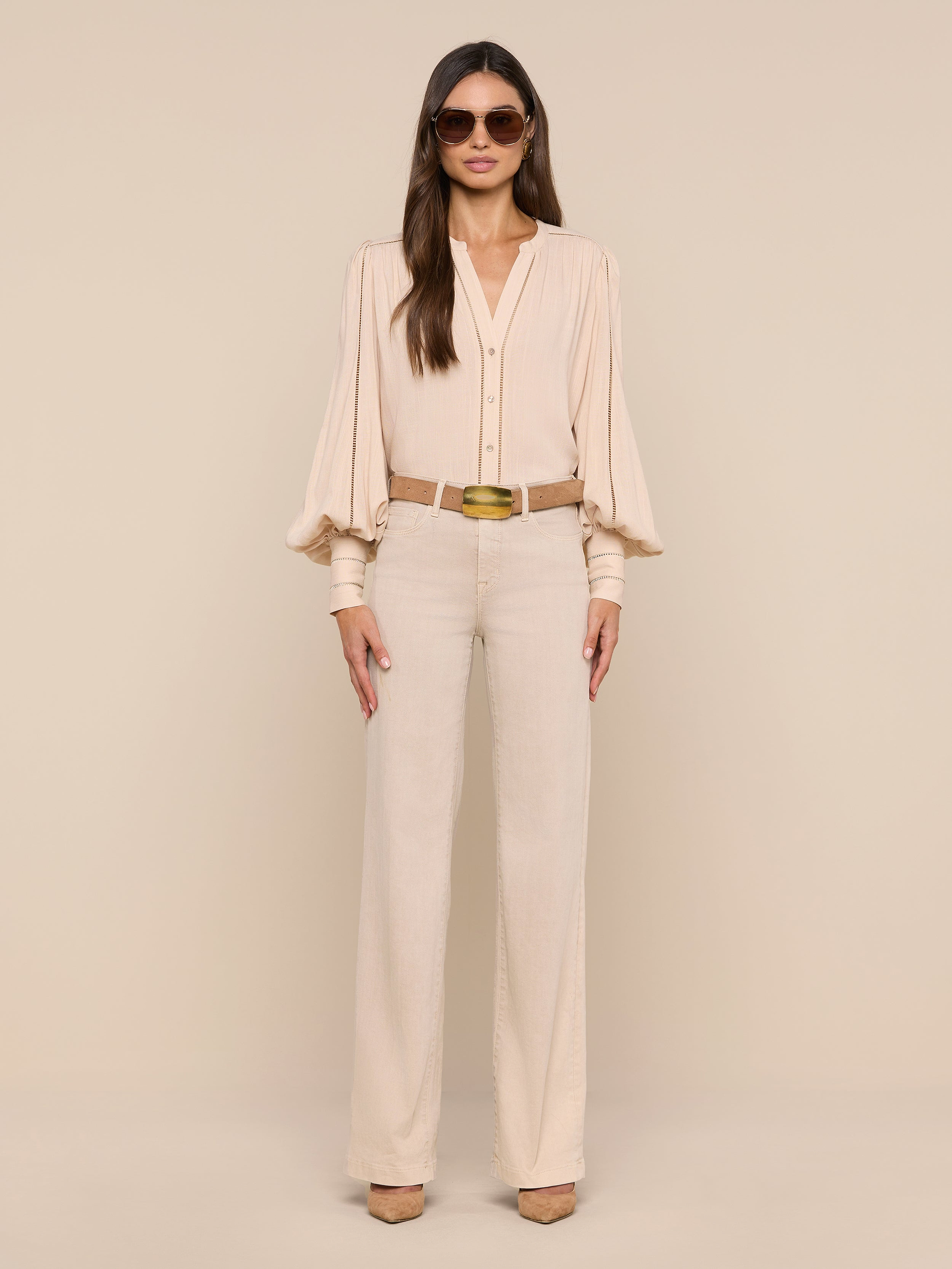 A woman stands confidently in a neutral studio, wearing the Kiera Blouse—a beige bohemian top with blouson sleeves—paired with matching high-waisted trousers, a brown belt, beige heels, and large sunglasses. Her long straight hair and poised stance complete the look.