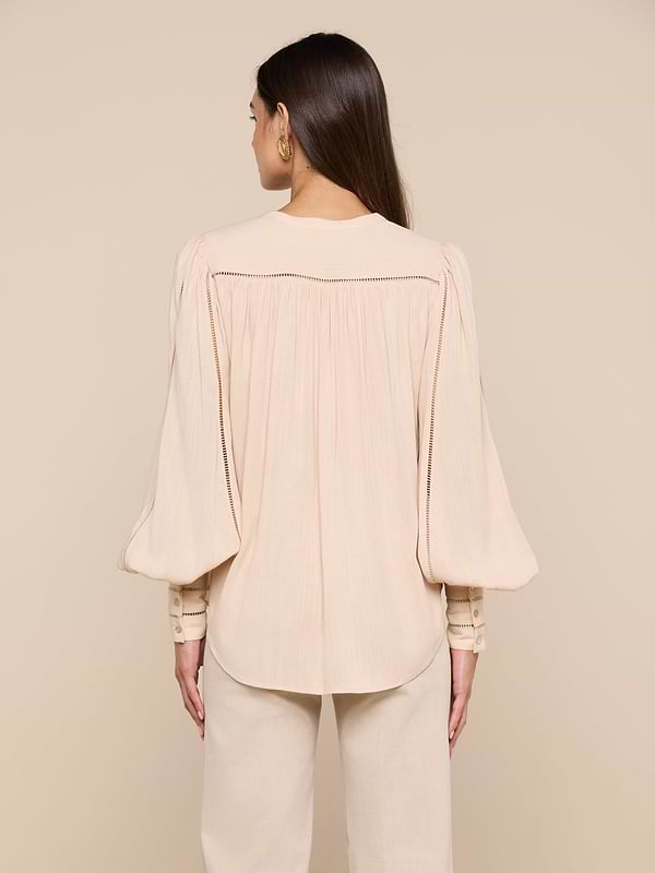 A woman stands with her back to the camera, wearing the Kiera Blouse—a loose, light beige bohemian top crafted from textured rayon with long blouson sleeves—paired with matching pants. The background is a plain off-white.