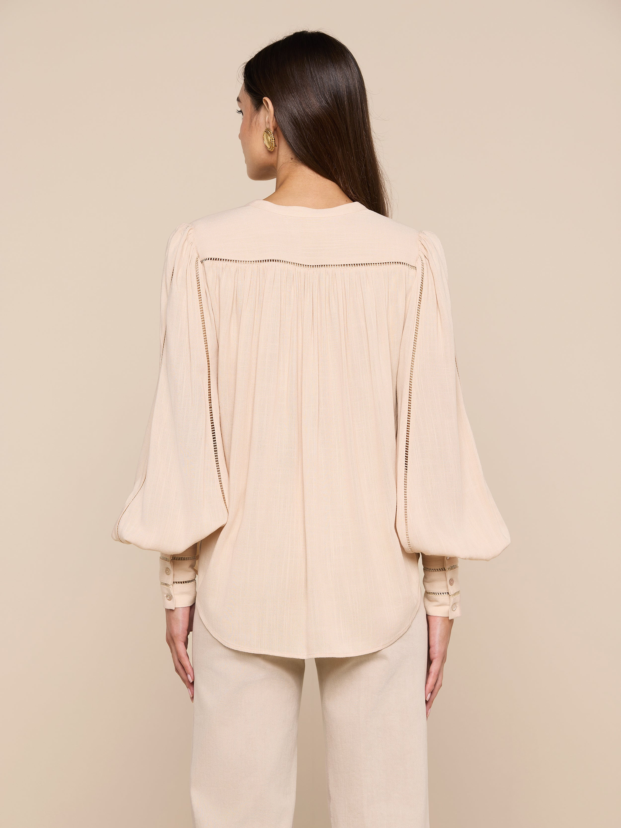 A woman stands with her back to the camera, wearing the Kiera Blouse—a loose, light beige bohemian top crafted from textured rayon with long blouson sleeves—paired with matching pants. The background is a plain off-white.