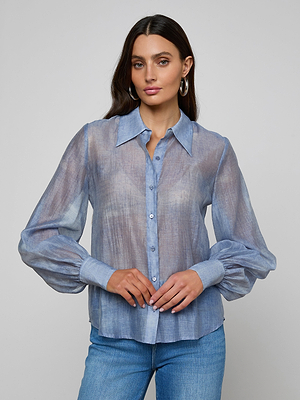 A woman with long dark hair wears the Jayleen Blouse—a light blue sheer chiffon top with blouson sleeves—paired with blue jeans, standing against a plain light gray background.
